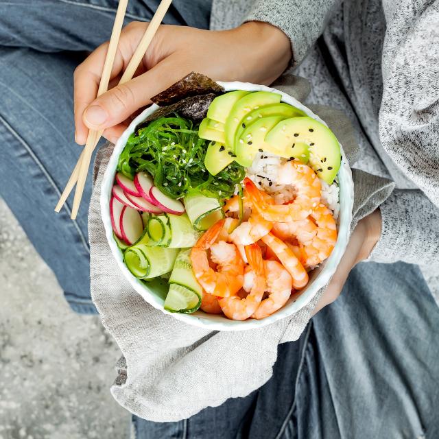 Girl in jeans holding shrimp poke bowl with seaweed, avocado, cucumber, radish, sesame seeds.